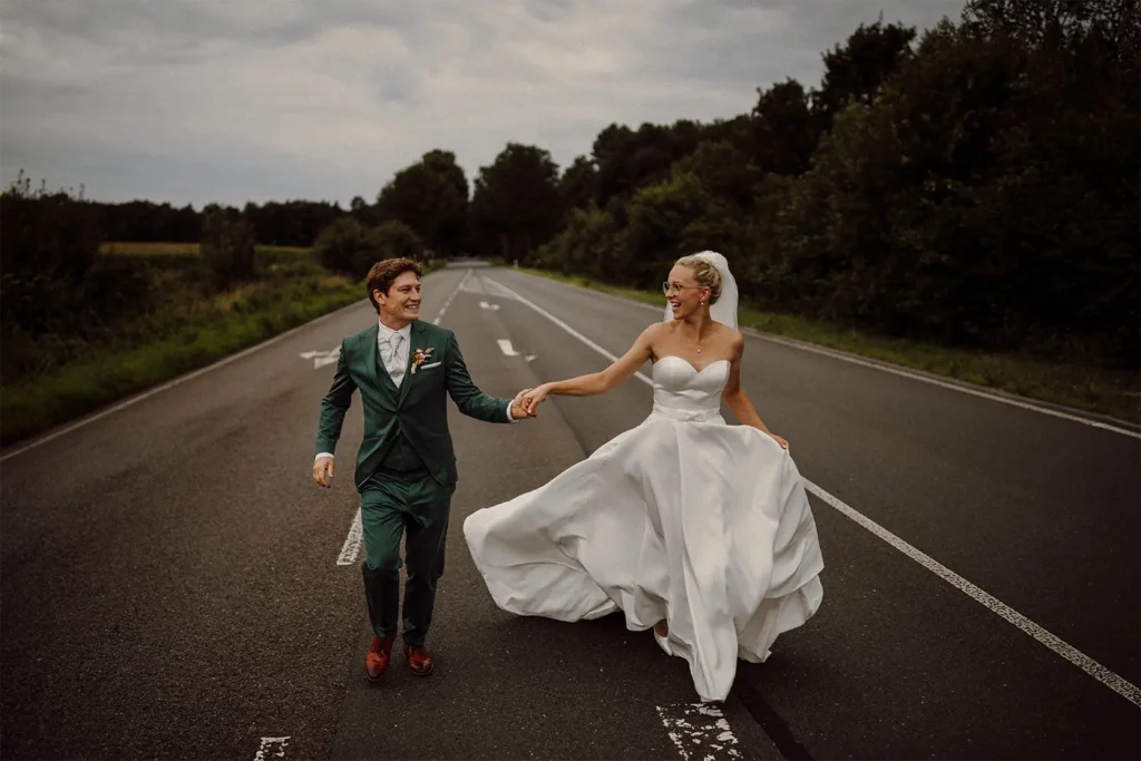 Fröhliches Brautpaar händehaltend auf leerer Landstraße, die Braut im weißen Hochzeitskleid und der Bräutigam in grünem Anzug.