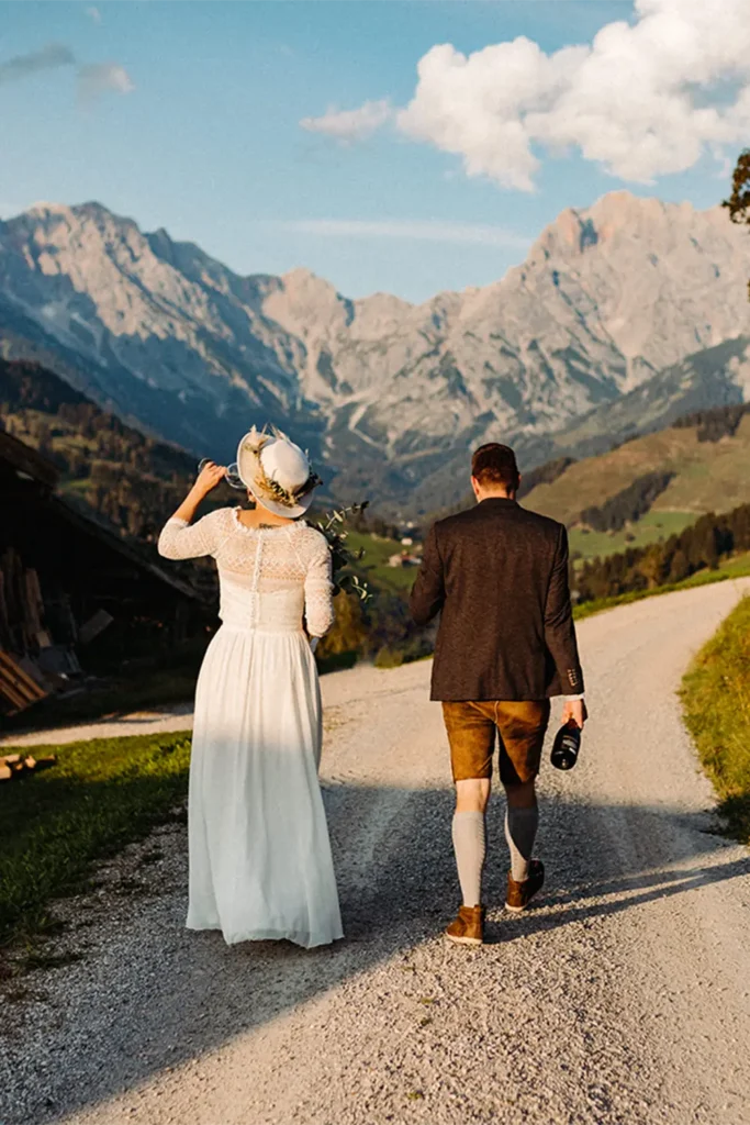 Rückenansicht eines Brautpaares in traditioneller Tracht, Hand in Hand gehend auf einem Weg mit malerischem Bergpanorama im Hintergrund.