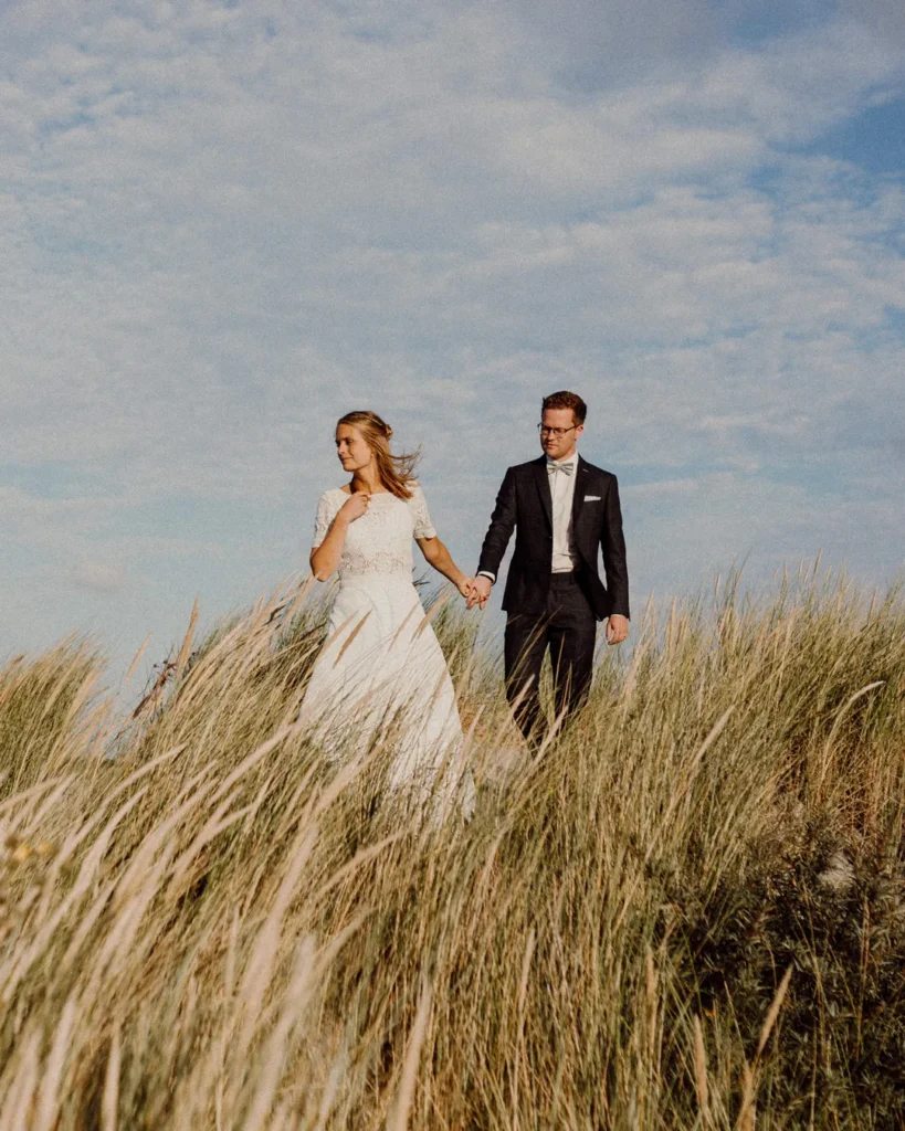 Brautpaar hand in hand spazierend durch hohes Gras mit blauem Himmel im Hintergrund, fotografiert von Björn Schreiber, Hochzeitsfotograf.